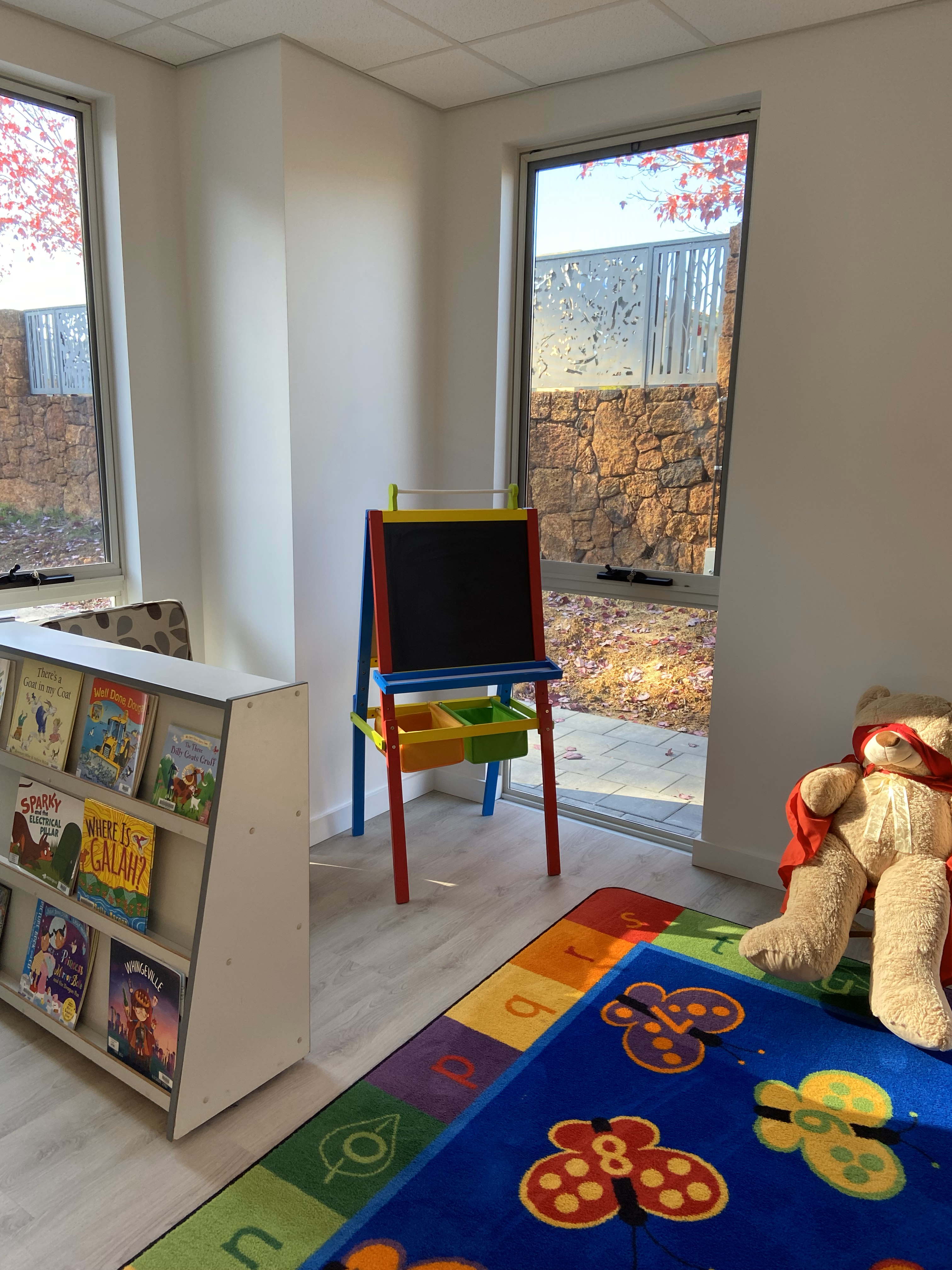 Interior photo of part of new Pemberton Library childrens corner, with bookshelf, colourful floor rug, art easel and teddy bear.